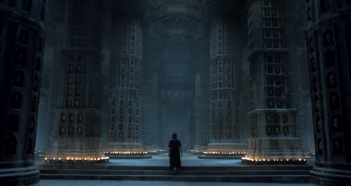 in a darkly candle lit hall, a woman walks up stairs, with columns covered in niches, each with a face
