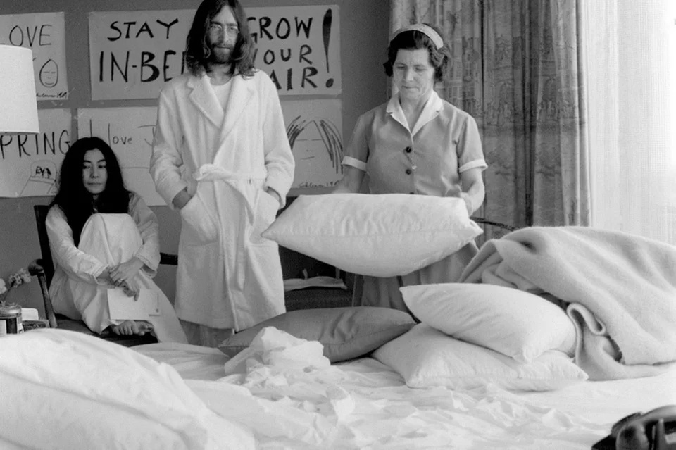 Black and white photo of a maid making up a bed, while John Lennon and Yoko Ono wait in their pajamas with protest signs on the wall